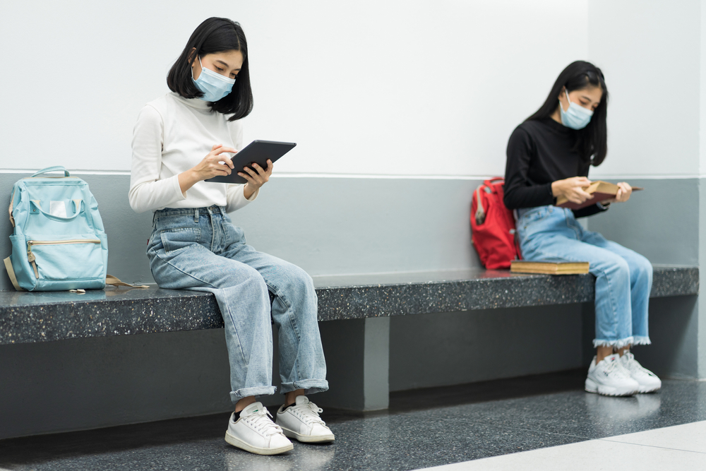 Two female college students wearing mask, reading book and learning while keeping social distance in university campus during COVID-19 pandemic